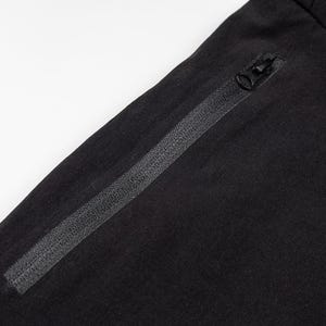 Pu&ograve; includere: Primo piano di pantaloni sportivi neri con tasca con cerniera e coulisse. I pantaloni sono realizzati in un tessuto scuro e morbido. La cerniera &egrave; nera e ha una piccola linguetta. La coulisse &egrave; anch'essa nera.
