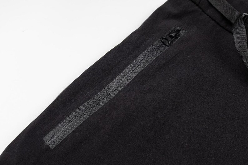 Pu&ograve; includere: Primo piano di pantaloni sportivi neri con tasca con cerniera e coulisse. I pantaloni sono realizzati in un tessuto scuro e morbido. La cerniera &egrave; nera e ha una piccola linguetta. La coulisse &egrave; anch'essa nera.