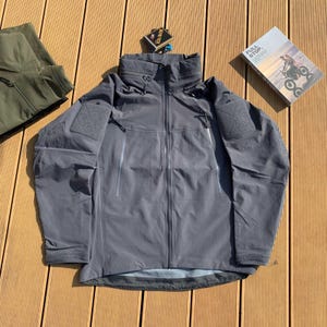 Op de afbeelding: Een grijze jas met capuchon, meerdere zakken en een ritssluiting. De jas heeft patches op de mouwen en een hoge kraag. Een label met de tekst "GORE-TEX" is bevestigd. Een boek met de tekst "FULL STOP" is zichtbaar.