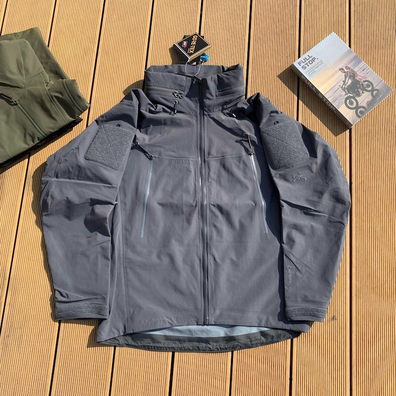 Op de afbeelding: Een grijze jas met capuchon, meerdere zakken en een ritssluiting. De jas heeft patches op de mouwen en een hoge kraag. Een label met de tekst "GORE-TEX" is bevestigd. Een boek met de tekst "FULL STOP" is zichtbaar.