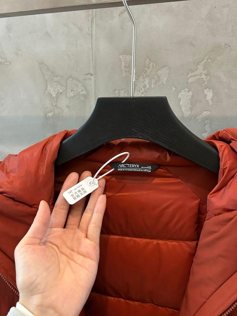 Op de afbeelding: Een roestkleurige Arc'teryx donsjas hangt aan een zwarte hanger. De jas heeft een gewatteerd ontwerp en een label met Chinese karakters. Het merklabel is zichtbaar. De jas is gemaakt van een glanzend, glad materiaal.