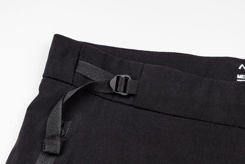 Pu&ograve; includere: Primo piano di pantaloni neri con fibbia e cinturino neri. I pantaloni sono realizzati in tessuto scuro e hanno un design minimalista. L'attenzione &egrave; focalizzata sulla cintura e sul cinturino funzionale.