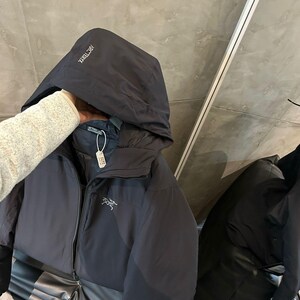 Op de afbeelding: Een zwarte jas met capuchon en ritssluiting. De jas heeft de merknaam "Arc'teryx" op de capuchon en een logo op de borst. De jas wordt op een zwarte ondergrond getoond.