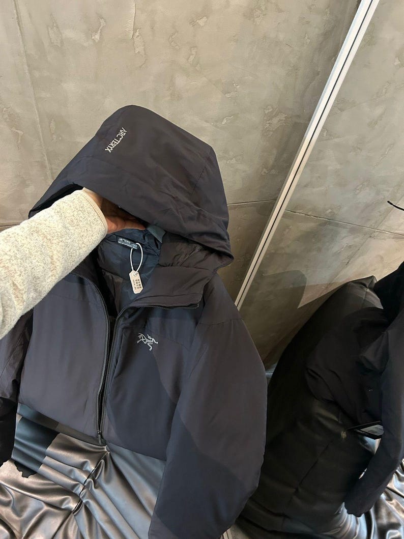 Op de afbeelding: Een zwarte jas met capuchon en ritssluiting. De jas heeft de merknaam "Arc'teryx" op de capuchon en een logo op de borst. De jas wordt op een zwarte ondergrond getoond.