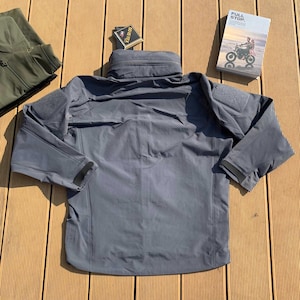 Op de afbeelding: Een grijze tactische jas met een hoge kraag en meerdere zakken. De jas heeft patches op de schouders en zwarte manchet details. Een klein boek met de woorden "FULL STOP" is zichtbaar.