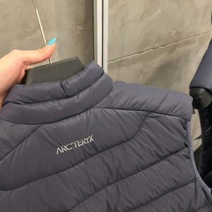 May include: A dark gray quilted vest with the brand name "ARC'TERYX" printed in white. The vest has a high collar and a diagonal quilted pattern. The vest is hanging on a black hanger.