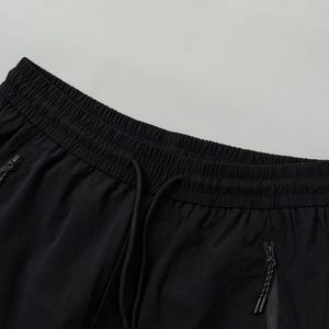 Pode incluir: Close-up de cal&ccedil;as de treino pretas. As cal&ccedil;as t&ecirc;m c&oacute;s com cord&atilde;o e dois bolsos com z&iacute;per. O tecido parece leve e ligeiramente texturizado. O fundo &eacute; cinza claro liso.