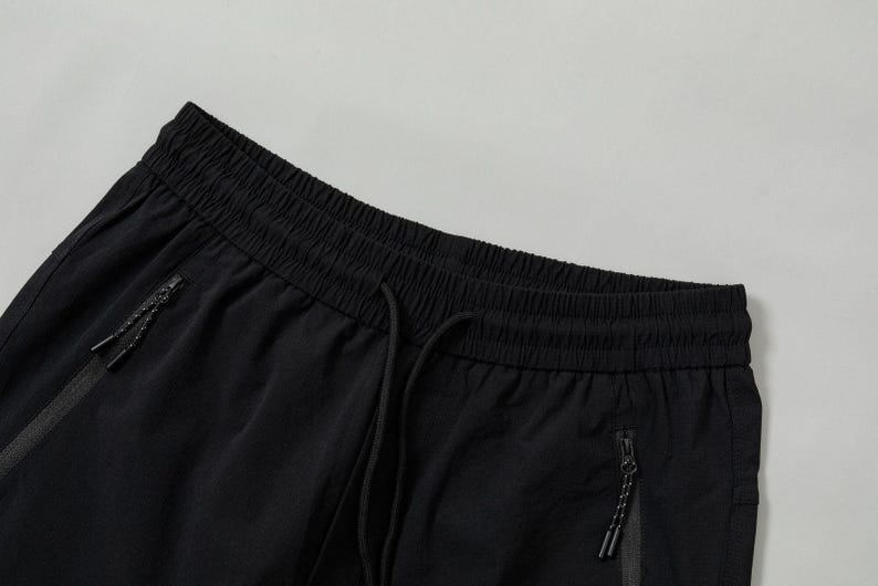 Pode incluir: Close-up de cal&ccedil;as de treino pretas. As cal&ccedil;as t&ecirc;m c&oacute;s com cord&atilde;o e dois bolsos com z&iacute;per. O tecido parece leve e ligeiramente texturizado. O fundo &eacute; cinza claro liso.