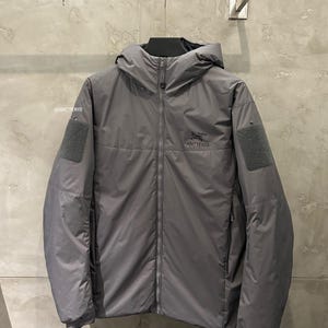 May include: A gray hooded jacket with a full-length zipper, featuring the brand name "Arc'teryx" on the chest. The jacket has two patches on the sleeves and a small orange pull cord. The jacket is hanging on a wall.