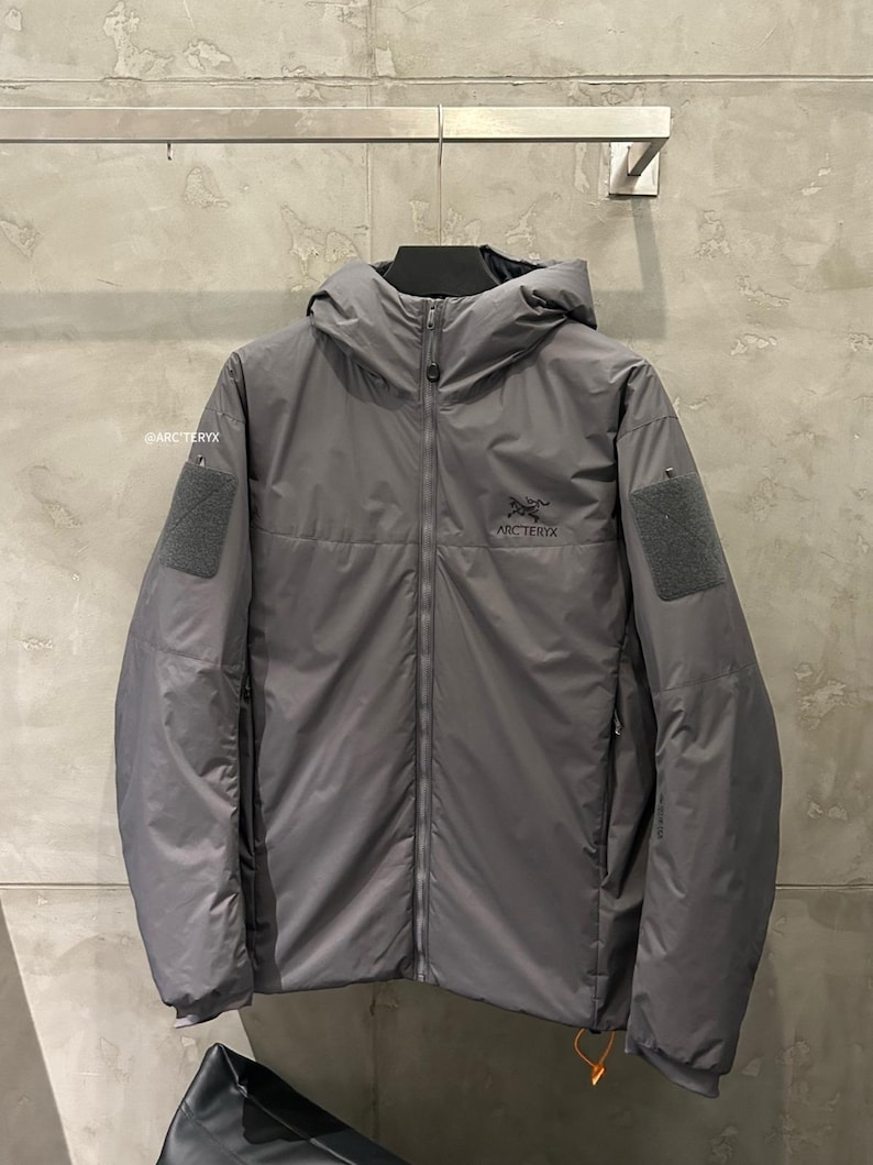 May include: A gray hooded jacket with a full-length zipper, featuring the brand name "Arc'teryx" on the chest. The jacket has two patches on the sleeves and a small orange pull cord. The jacket is hanging on a wall.