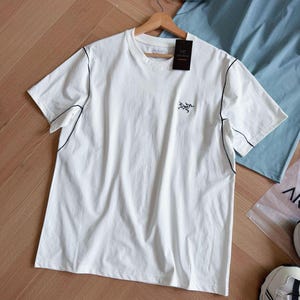 May include: A white t-shirt with black stitching along the seams and a small logo on the chest. The shirt is hanging on a wooden hanger with a black tag attached. A light blue shirt is visible in the background.