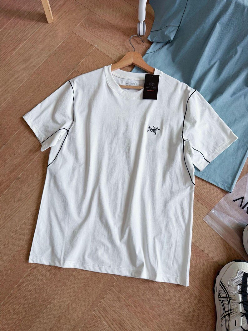 May include: A white t-shirt with black stitching along the seams and a small logo on the chest. The shirt is hanging on a wooden hanger with a black tag attached. A light blue shirt is visible in the background.