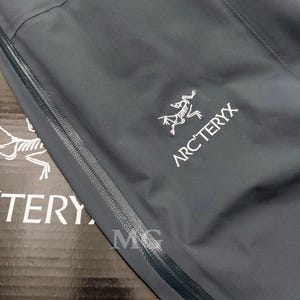 K&ouml;nnte beinhalten: Nahaufnahme einer dunkelgrauen Jacke mit Rei&szlig;verschluss und dem wei&szlig;en Arc'teryx-Logo. Das Logo zeigt eine stilisierte, dinosaurierartige Kreatur. Die Jacke scheint aus einem glatten, wasserabweisenden Material zu bestehen.