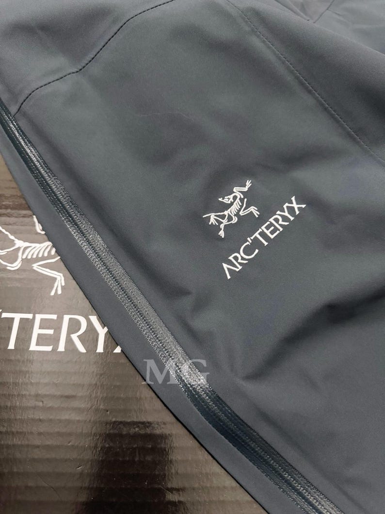 K&ouml;nnte beinhalten: Nahaufnahme einer dunkelgrauen Jacke mit Rei&szlig;verschluss und dem wei&szlig;en Arc'teryx-Logo. Das Logo zeigt eine stilisierte, dinosaurierartige Kreatur. Die Jacke scheint aus einem glatten, wasserabweisenden Material zu bestehen.