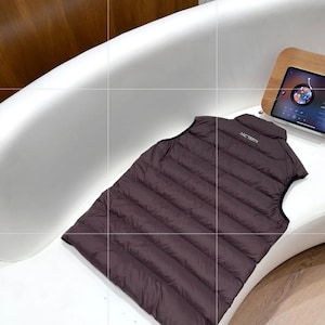 May include: A dark brown, quilted vest with the brand name "Arc'teryx" printed on the collar. The vest is sleeveless and appears to be made of a lightweight, possibly down-filled material. It is lying on a white curved surface.
