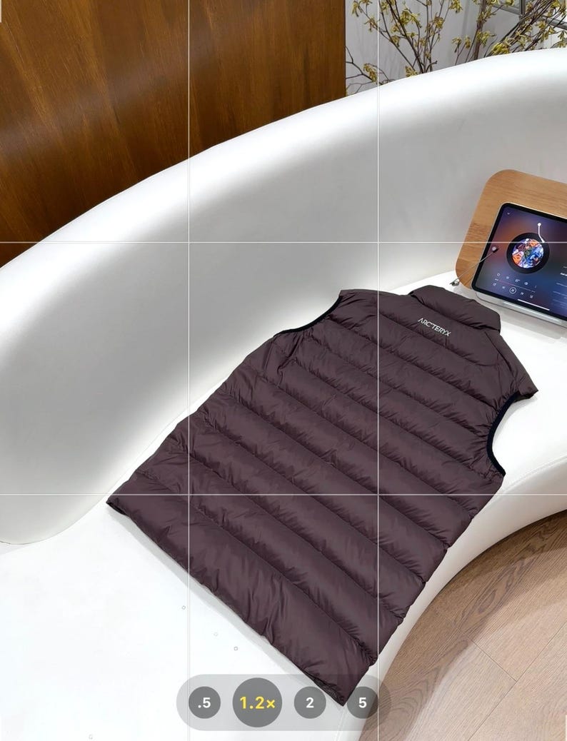 May include: A dark brown, quilted vest with the brand name "Arc'teryx" printed on the collar. The vest is sleeveless and appears to be made of a lightweight, possibly down-filled material. It is lying on a white curved surface.