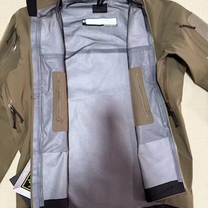 Op de afbeelding: Open olijfgroene jas met een volledige ritssluiting, meerdere zakken en een zwarte kraag. De jas heeft een grijze binnenkant met een label "Arc'teryx". De jas is ontworpen voor buitenactiviteiten.