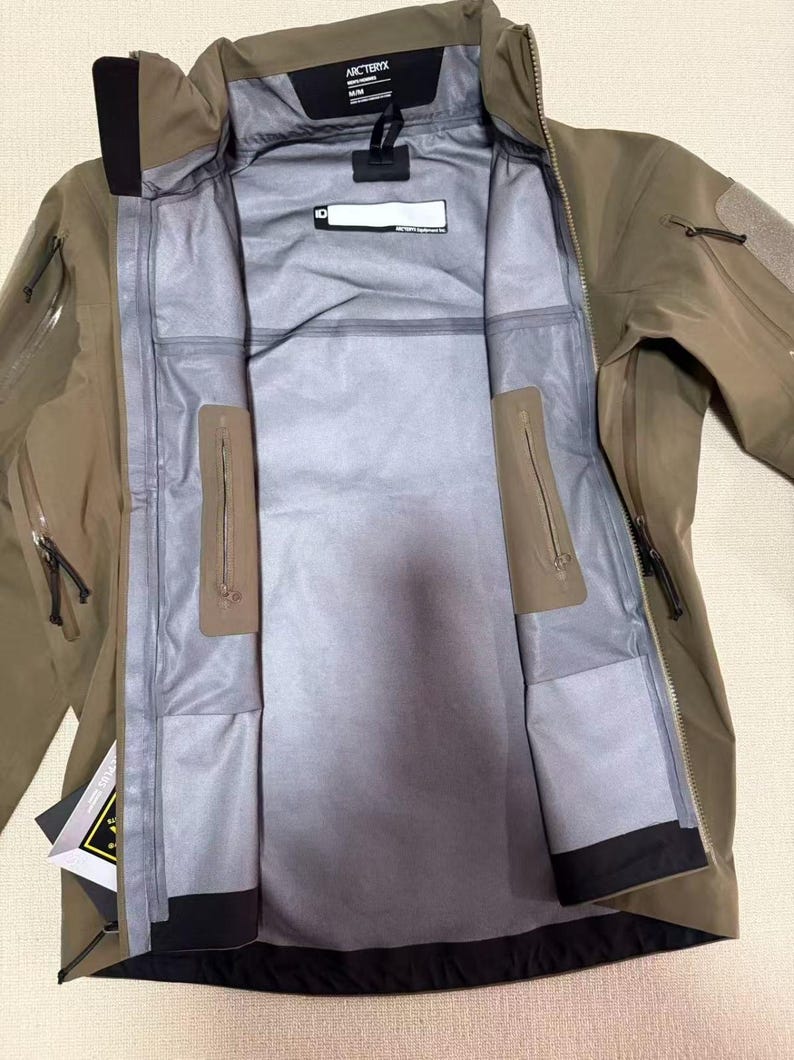 Op de afbeelding: Open olijfgroene jas met een volledige ritssluiting, meerdere zakken en een zwarte kraag. De jas heeft een grijze binnenkant met een label "Arc'teryx". De jas is ontworpen voor buitenactiviteiten.