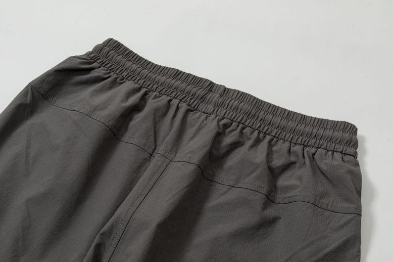 Pode incluir: Close-up da cintura e da parte superior de cal&ccedil;as esportivas cinza escuro. A cintura el&aacute;stica &eacute; franzida e costurada, com um canal para o cord&atilde;o. O tecido parece leve e ligeiramente texturizado, com costuras verticais.