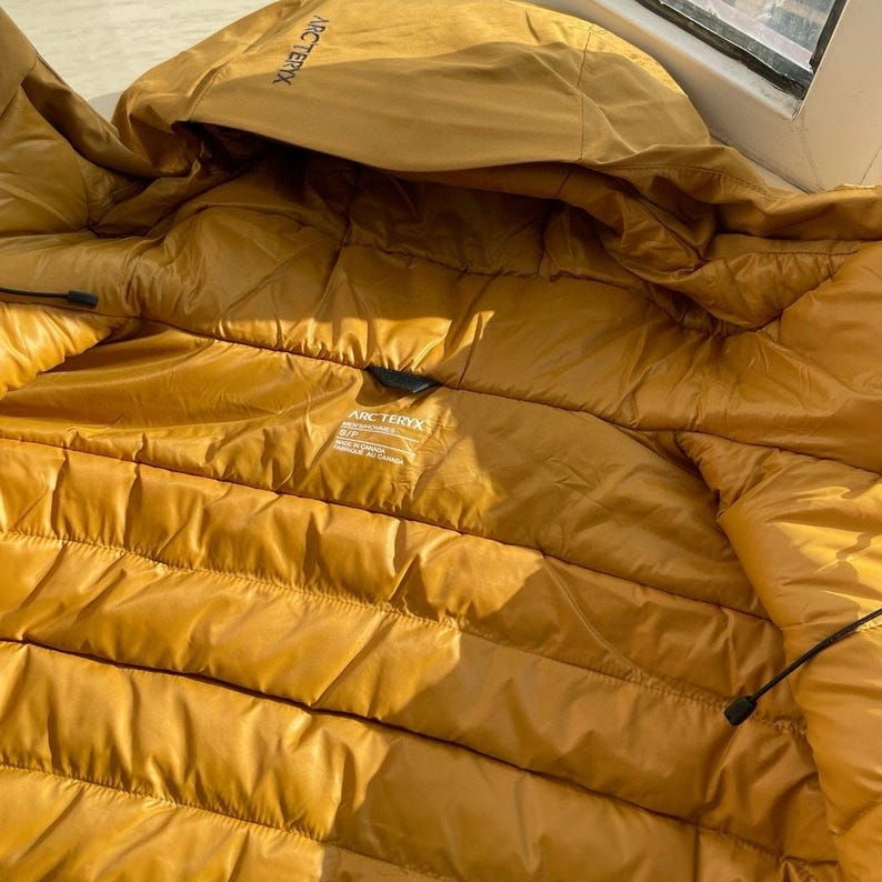May include: Mustard-coloured puffer jacket with a hood. The jacket has a quilted design and the brand name "Arc'teryx" is visible on the hood and inside label. The jacket is made of a shiny, water-resistant material.