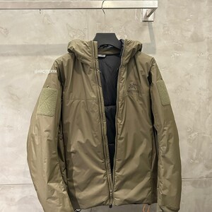 May include: Olive green hooded jacket with a full-length zipper. The jacket features a black interior lining, a small patch on the left sleeve, and the brand name "ARC'TERYX" printed on the chest. The jacket is hanging on a metal bar.
