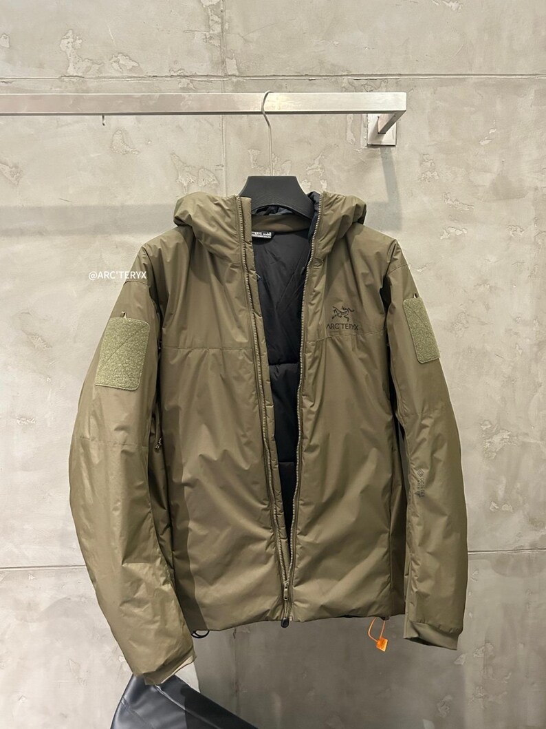 May include: Olive green hooded jacket with a full-length zipper. The jacket features a black interior lining, a small patch on the left sleeve, and the brand name "ARC'TERYX" printed on the chest. The jacket is hanging on a metal bar.