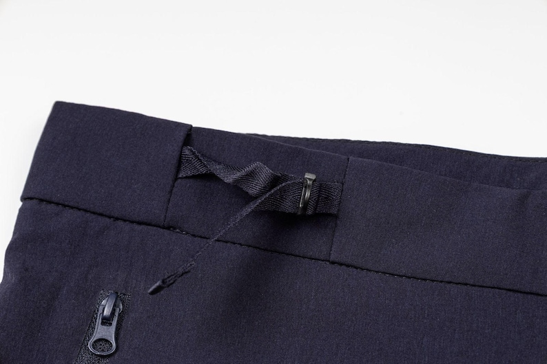 Pu&ograve; includere: Primo piano di pantaloni sportivi blu navy. La cintura presenta un cordino e una chiusura in metallo. Una tasca con cerniera &egrave; visibile sul lato sinistro. Il tessuto ha una trama sottile.