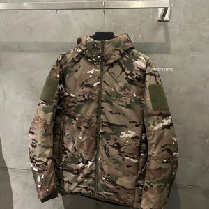 May include: A camouflage jacket with a hood, zipper closure, and two green patches on the sleeves. The jacket features a mix of green, brown, and white patterns. The brand name "ARC'TERYX" is visible on the chest.