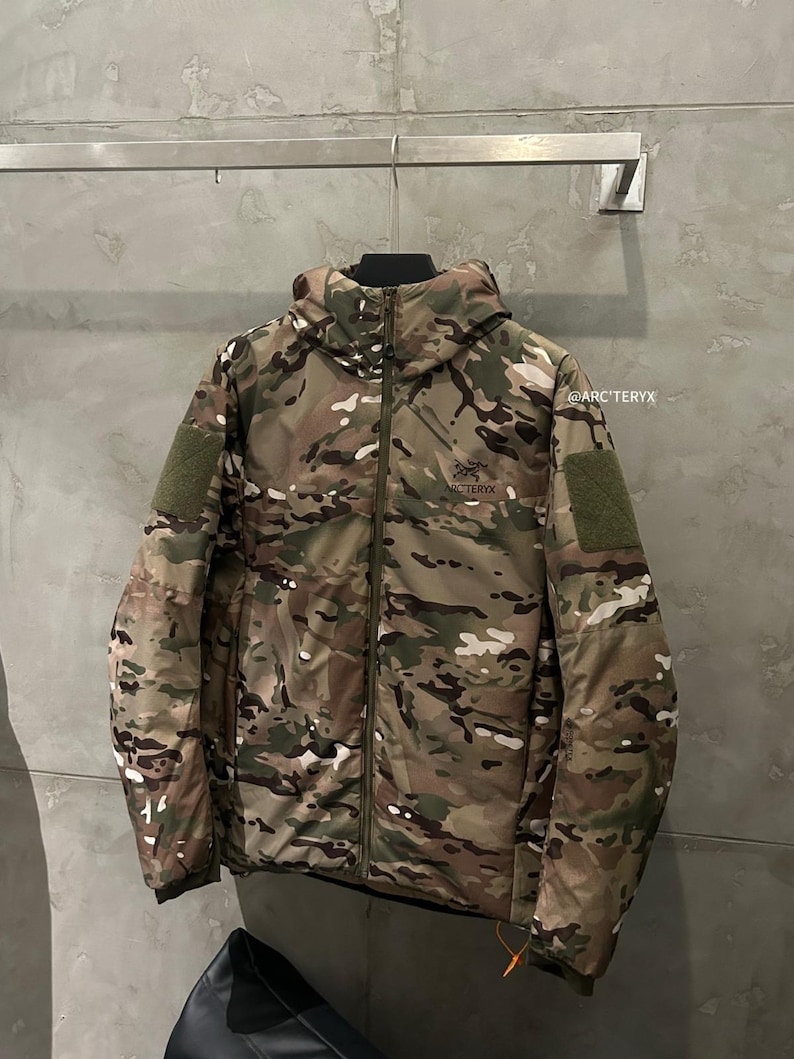 May include: A camouflage jacket with a hood, zipper closure, and two green patches on the sleeves. The jacket features a mix of green, brown, and white patterns. The brand name "ARC'TERYX" is visible on the chest.
