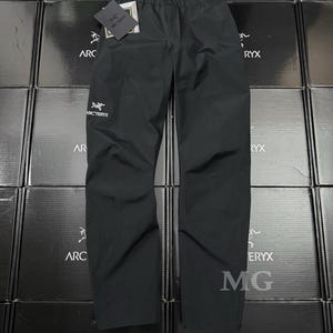 K&ouml;nnte beinhalten: Schwarze wasserdichte Hose mit dem Arc'teryx-Logo und einem Etikett. Die Hose wird auf einem Stapel von Kartons mit dem Arc'teryx-Logo pr&auml;sentiert. Der Text "Beta 全拉鏈衝鋒褲" ist auf den Kartons zu sehen.