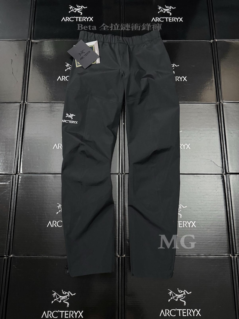 K&ouml;nnte beinhalten: Schwarze wasserdichte Hose mit dem Arc'teryx-Logo und einem Etikett. Die Hose wird auf einem Stapel von Kartons mit dem Arc'teryx-Logo pr&auml;sentiert. Der Text "Beta 全拉鏈衝鋒褲" ist auf den Kartons zu sehen.