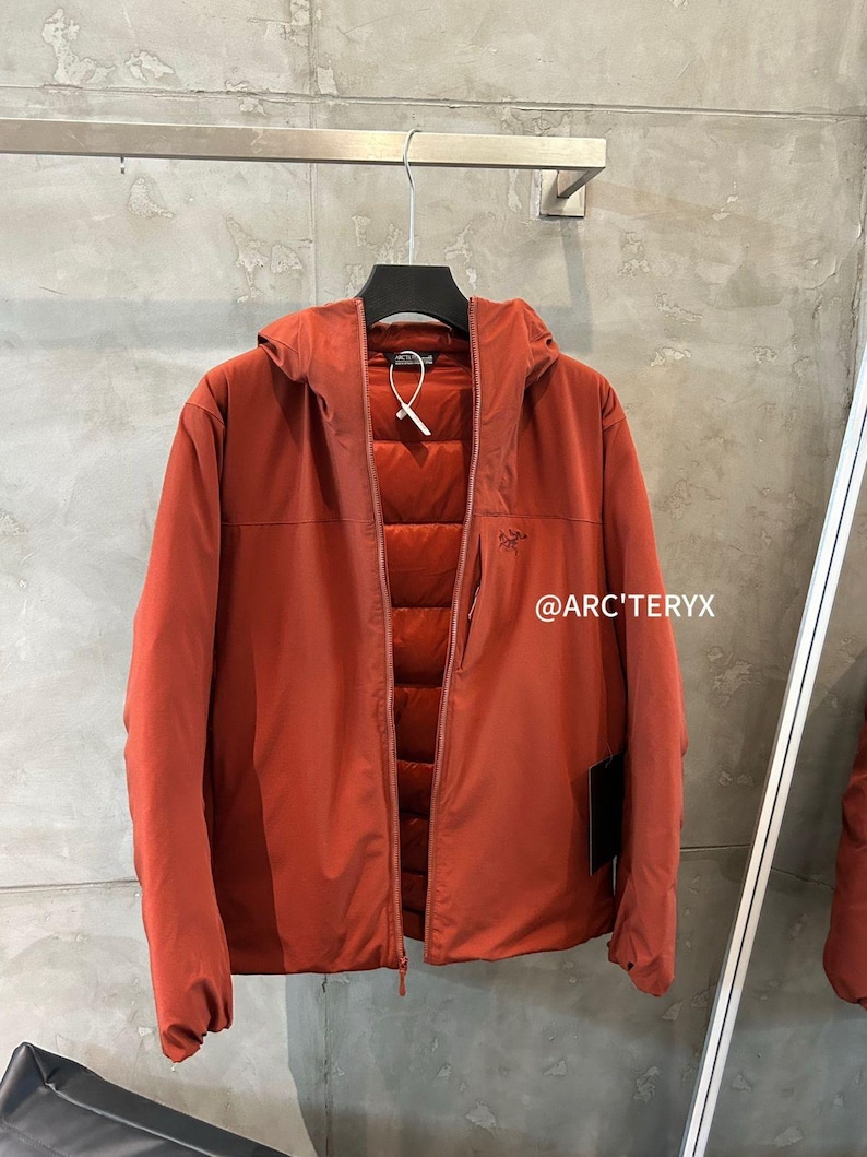 Op de afbeelding: Een roestkleurige jas met capuchon en ritssluiting, hangend aan een zwarte hanger. De jas heeft een gewatteerde binnenkant en de tekst "@ARC'TERYX" is zichtbaar aan de rechterkant.