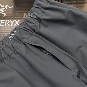K&ouml;nnte beinhalten: Nahaufnahme einer dunkelgrauen Sporthose mit Rei&szlig;verschlusstasche. Die Hose hat einen elastischen Bund und der Markenname "Arc'teryx" ist sichtbar. Der Stoff scheint ein leichtes, strapazierf&auml;higes Material zu sein.