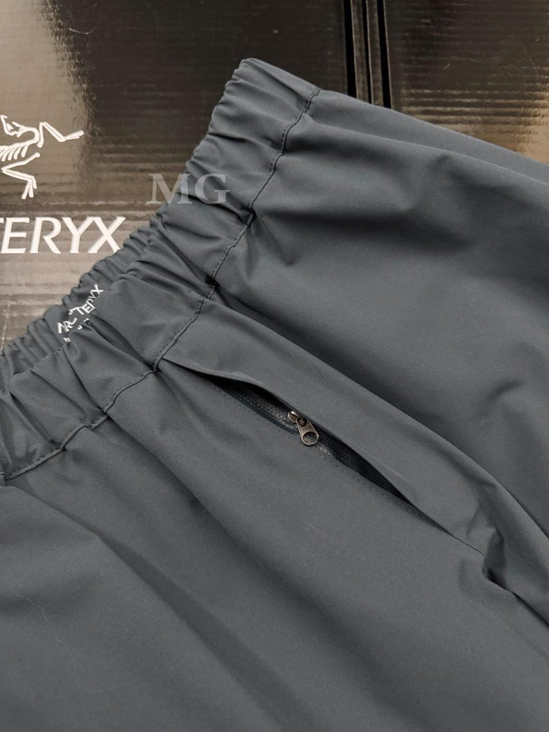 K&ouml;nnte beinhalten: Nahaufnahme einer dunkelgrauen Sporthose mit Rei&szlig;verschlusstasche. Die Hose hat einen elastischen Bund und der Markenname "Arc'teryx" ist sichtbar. Der Stoff scheint ein leichtes, strapazierf&auml;higes Material zu sein.
