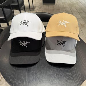 Puede incluir: Una colecci&oacute;n de gorras de b&eacute;isbol en varios colores, incluyendo blanco, negro, beige y gris. Cada gorra presenta un logotipo blanco bordado en la parte delantera. Las gorras se exhiben sobre una mesa redonda y oscura.