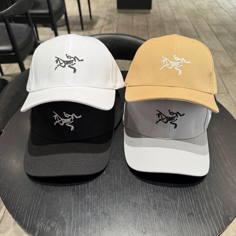 Puede incluir: Una colecci&oacute;n de gorras de b&eacute;isbol en varios colores, incluyendo blanco, negro, beige y gris. Cada gorra presenta un logotipo blanco bordado en la parte delantera. Las gorras se exhiben sobre una mesa redonda y oscura.