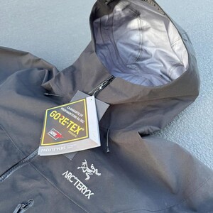 May include: Black hooded jacket with a zipper closure and the Arc'teryx logo. The jacket features a Gore-Tex label, indicating waterproof and breathable fabric. The hood is open, revealing a light gray interior.