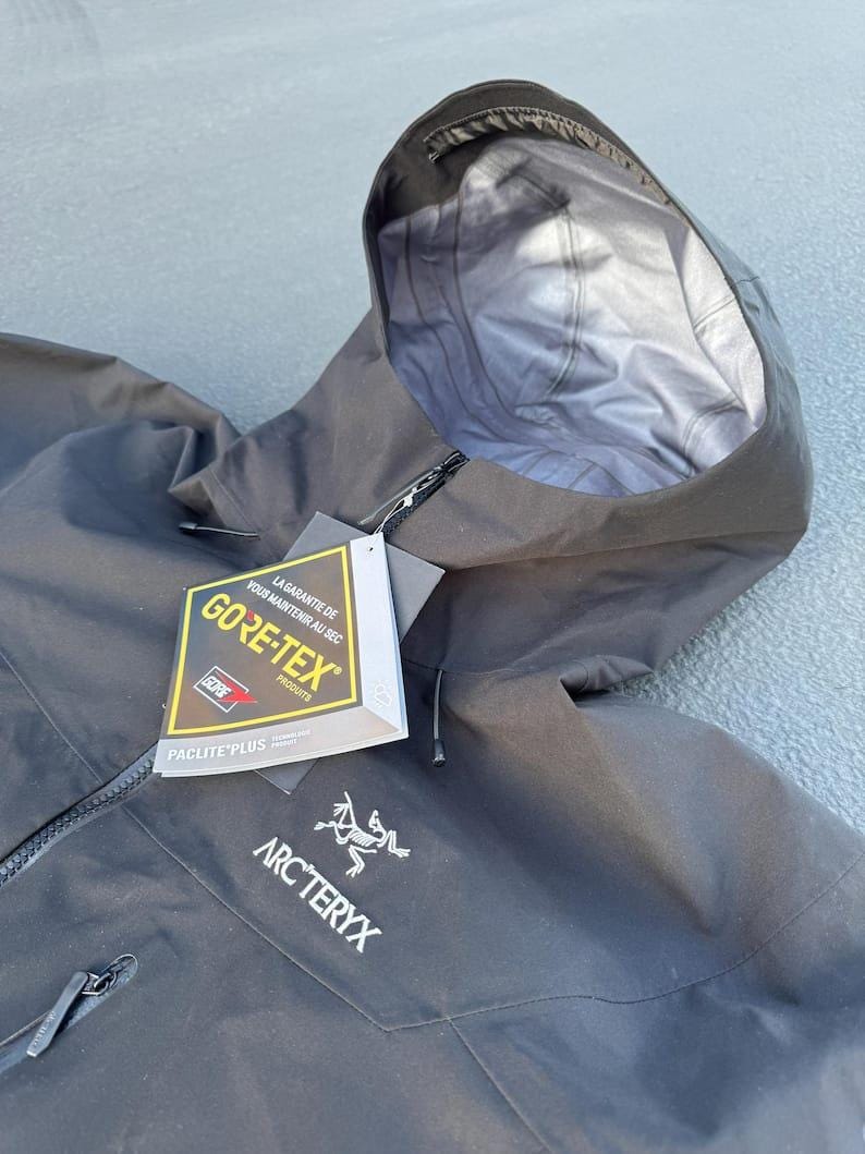 May include: Black hooded jacket with a zipper closure and the Arc'teryx logo. The jacket features a Gore-Tex label, indicating waterproof and breathable fabric. The hood is open, revealing a light gray interior.