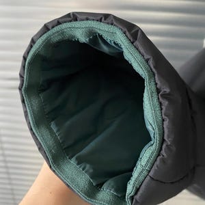 Op de afbeelding: Close-up van een zwarte pufferjackmouw. De binnenvoering is donkerblauwgroen en de manchet is afgezet met een blauwgroene stof. De jas lijkt gemaakt te zijn van een gewatteerd materiaal.
