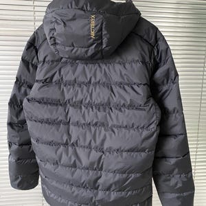 Op de afbeelding: Zwarte pufferjack met capuchon, hangend aan een zwarte hanger. De jas heeft horizontale gewatteerde lijnen en de merknaam "ARCTERYX" is in goud op de capuchon gedrukt. Ontworpen voor koud weer.