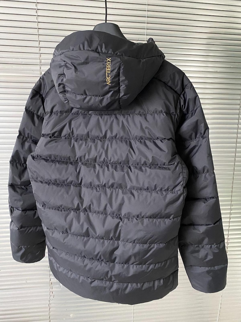 Op de afbeelding: Zwarte pufferjack met capuchon, hangend aan een zwarte hanger. De jas heeft horizontale gewatteerde lijnen en de merknaam "ARCTERYX" is in goud op de capuchon gedrukt. Ontworpen voor koud weer.