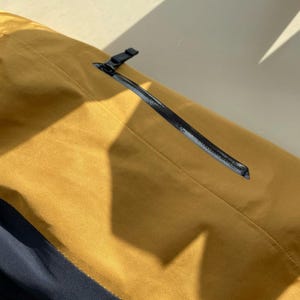 May include: Close-up of a mustard-coloured jacket with a black zip and a small black pull tab. The jacket has a subtle sheen and a seam running down the front. The bottom portion of the jacket is navy blue.