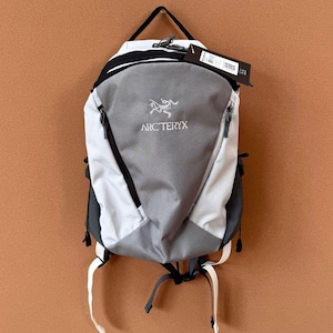 May include: A gray, white, and black backpack with the Arc'teryx logo. The backpack has a top handle, two side pockets, and adjustable straps. A tag hangs from the top of the backpack.