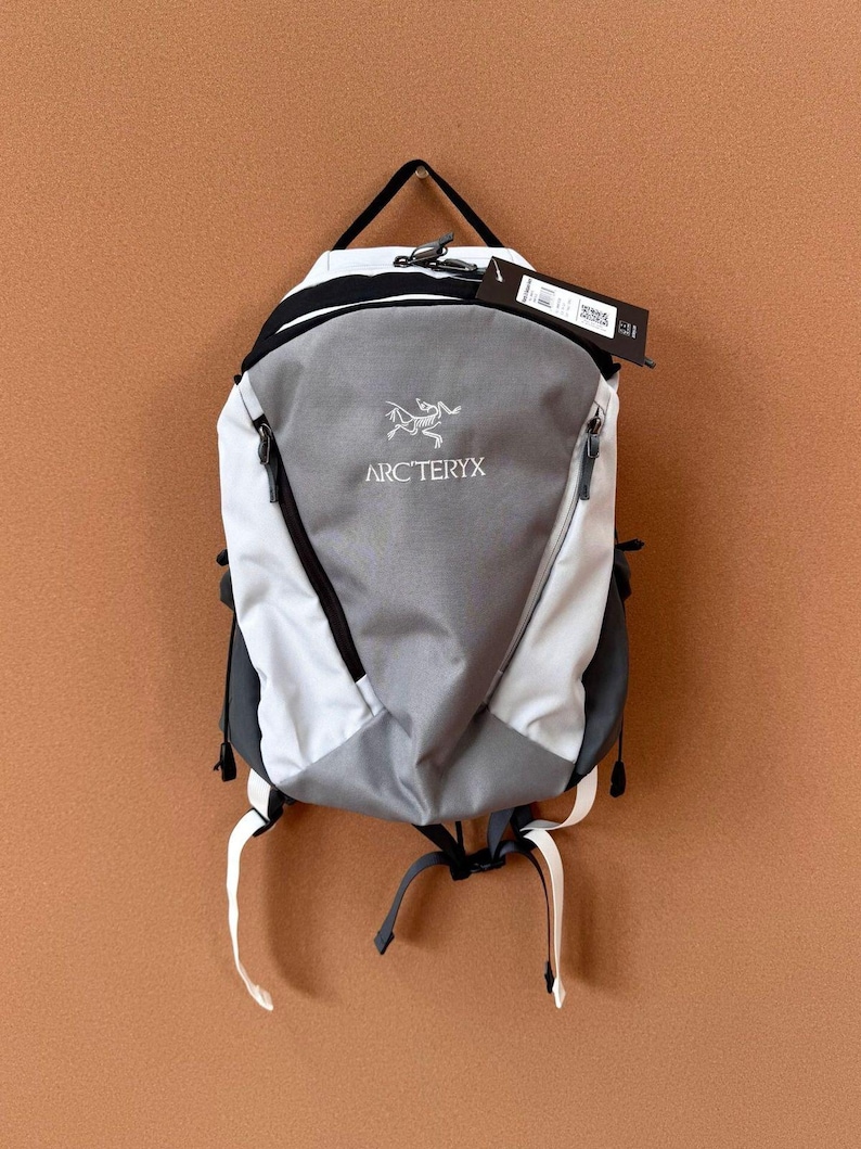 May include: A gray, white, and black backpack with the Arc'teryx logo. The backpack has a top handle, two side pockets, and adjustable straps. A tag hangs from the top of the backpack.