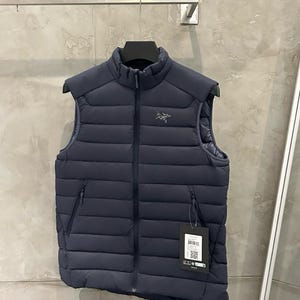 May include: Navy blue puffer vest with a high collar and a full-length zipper. The vest has two side pockets and a small logo on the chest. A tag hangs from the zipper. The vest is hanging on a metal bar.