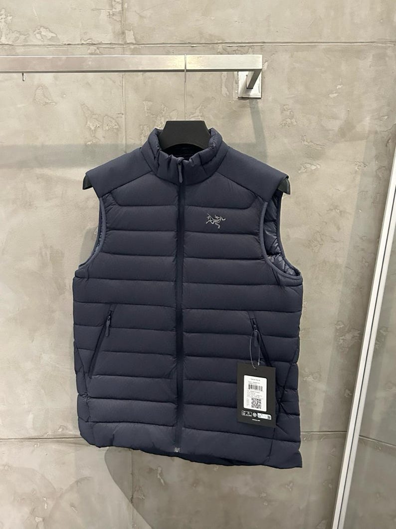 May include: Navy blue puffer vest with a high collar and a full-length zipper. The vest has two side pockets and a small logo on the chest. A tag hangs from the zipper. The vest is hanging on a metal bar.