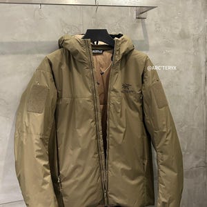 May include: A tan hooded jacket with a full-length zipper and two patch pockets. The jacket has the brand name "Arc'teryx" printed on the chest and the text "@ARC'TERYX" on the right sleeve.