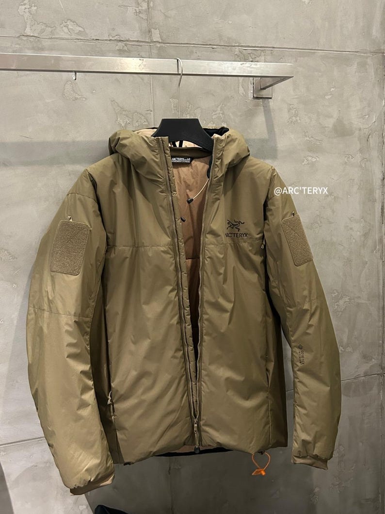 May include: A tan hooded jacket with a full-length zipper and two patch pockets. The jacket has the brand name "Arc'teryx" printed on the chest and the text "@ARC'TERYX" on the right sleeve.