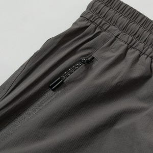 Pode incluir: Close-up de cal&ccedil;&otilde;es desportivos cinzentos com cintura com cord&atilde;o e bolso com z&iacute;per. Os cal&ccedil;&otilde;es s&atilde;o feitos de um tecido leve e texturizado. O puxador do z&iacute;per &eacute; preto com um padr&atilde;o de pontos brancos.