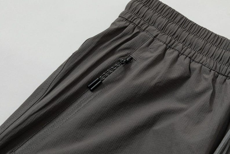 Pode incluir: Close-up de cal&ccedil;&otilde;es desportivos cinzentos com cintura com cord&atilde;o e bolso com z&iacute;per. Os cal&ccedil;&otilde;es s&atilde;o feitos de um tecido leve e texturizado. O puxador do z&iacute;per &eacute; preto com um padr&atilde;o de pontos brancos.